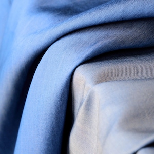 May include: Close-up of folded blue fabric with varying shades, from light to deep blue. The fabric has a smooth texture and appears to be a lightweight material, possibly suitable for clothing or home decor.