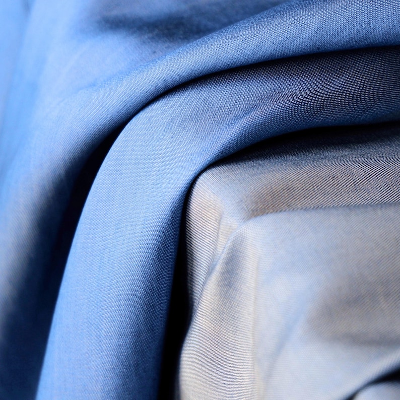 May include: Close-up of folded blue fabric with varying shades, from light to deep blue. The fabric has a smooth texture and appears to be a lightweight material, possibly suitable for clothing or home decor.