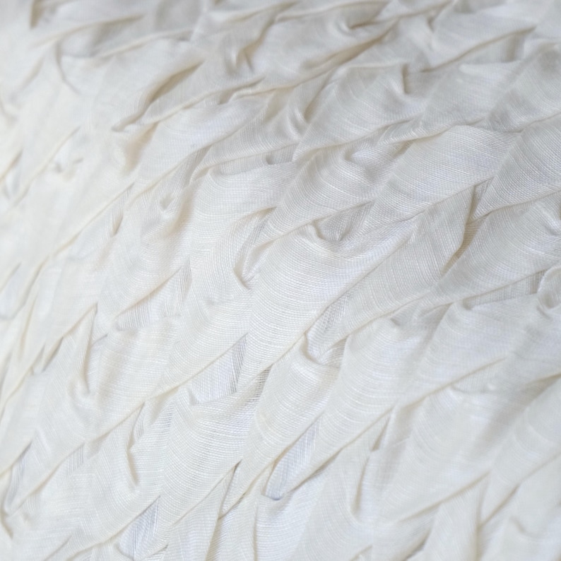 May include: Close-up of a white fabric with a textured, layered design. The fabric appears to be made of a soft material, with a rippled, wave-like pattern. The folds create shadows and depth, enhancing the visual texture.