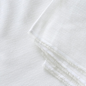 May include: A close-up shot of a white herringbone-patterned fabric. The textile is folded, showing multiple layers. The edges of the fabric have a fringed detail. The overall look is clean and simple.