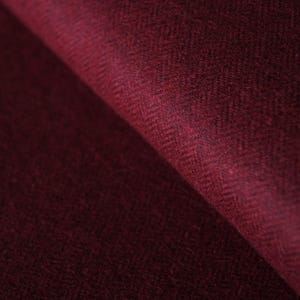 Puede incluir: Una tela de color burdeos profundo con un sutil patrón de espiga tejido. El material muestra una textura suave y ligeramente difusa con un pliegue diagonal que crea un degradado de luz y sombra sobre la rica superficie roja oscura.
