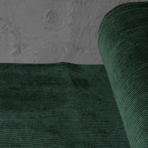 May include: A close-up of a roll of dark green corduroy fabric. The fabric is a deep, forest green color with a ribbed texture. The fabric is partially rolled, revealing the texture and color.