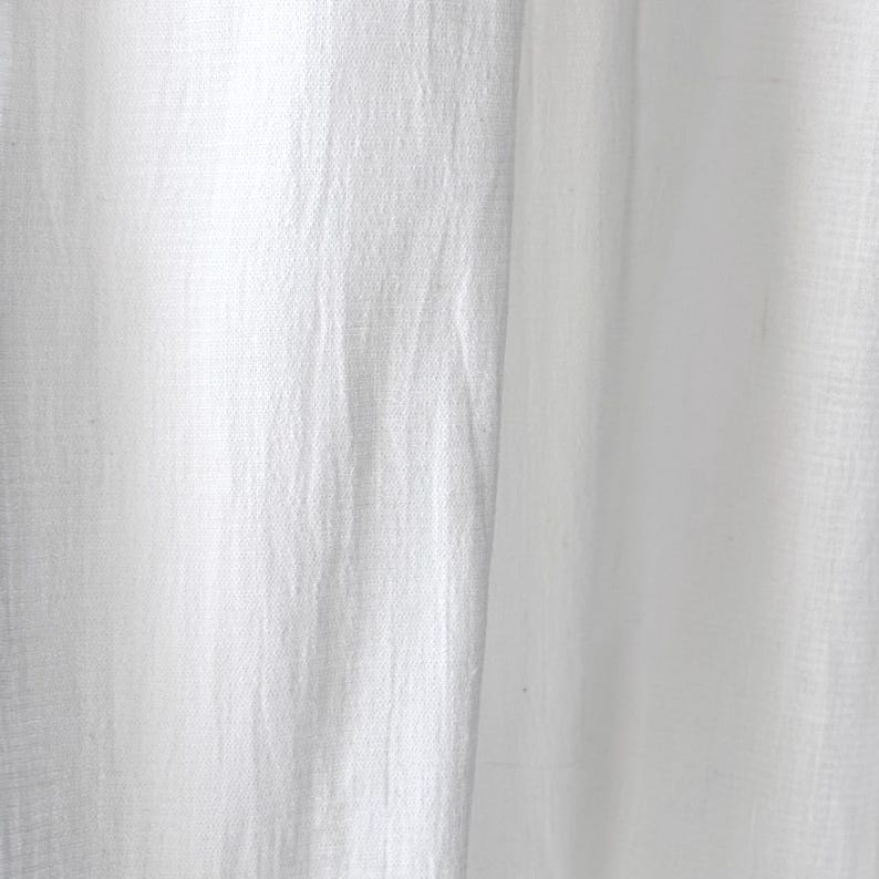 May include: Close-up of white fabric with a textured appearance. The fabric has a slightly wrinkled look, with vertical folds and subtle variations in light and shadow. The material appears to be lightweight and possibly linen or cotton.