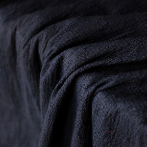 May include: Close-up of a dark navy blue fabric with a textured weave. The fabric is draped, creating soft folds and shadows. The material appears to be linen or a similar natural fiber, suitable for clothing or home decor.