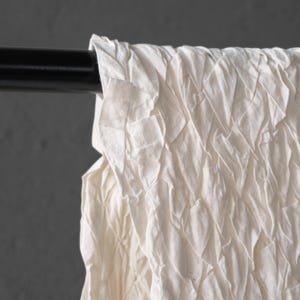 May include: Close-up of a white, textured fabric curtain hanging from a black rod. The fabric has a layered, diamond-like pattern, creating a soft, flowing appearance. The background is a solid, neutral gray.