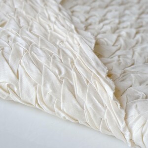 May include: Close-up of off-white fabric with a textured, woven design. The fabric is folded, revealing the intricate pattern of the weave. The edges of the fabric are unfinished, showing the raw texture of the material.