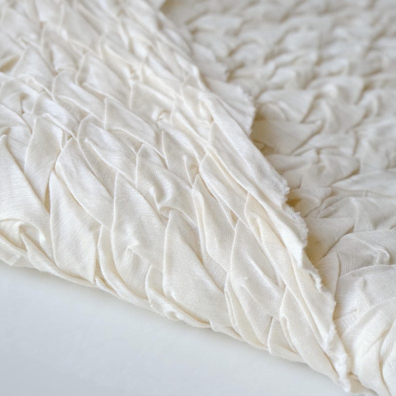 May include: Close-up of off-white fabric with a textured, woven design. The fabric is folded, revealing the intricate pattern of the weave. The edges of the fabric are unfinished, showing the raw texture of the material.