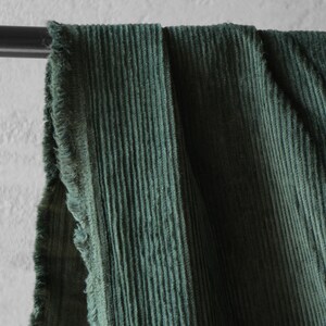 May include: Close-up of a section of green corduroy fabric. The fabric is draped over a black metal rod, with a frayed edge visible on the left side. The corduroy has vertical ridges and a soft texture.