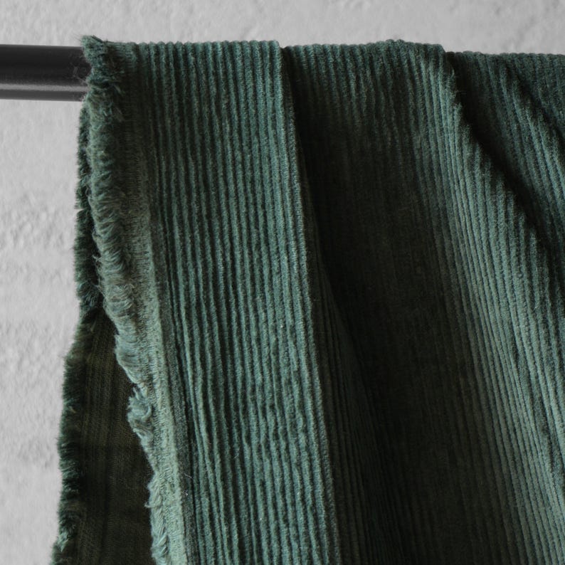 May include: Close-up of a section of green corduroy fabric. The fabric is draped over a black metal rod, with a frayed edge visible on the left side. The corduroy has vertical ridges and a soft texture.
