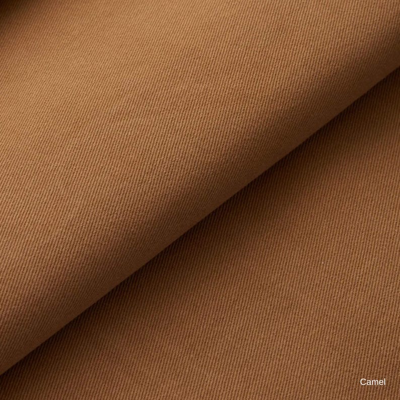 K&ouml;nnte beinhalten: Ein einfarbiger, kamelfarbener Stoff mit einem ausgepr&auml;gten diagonalen K&ouml;perbindungsmuster. Das Textil ist umgeschlagen, um die Textur und Tiefe des Gewebes zu zeigen. Das Wort Camel ist in der unteren rechten Ecke des Bildes aufgedruckt.