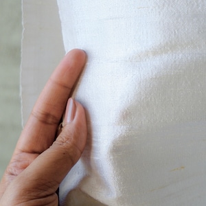 May include: Close-up of a piece of off-white fabric with a subtle sheen. The fabric has a textured surface and appears to be held by a hand. The fabric's edges are slightly frayed, suggesting it may be a cut piece of material.