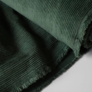 May include: Close-up of a roll of dark green corduroy fabric. The fabric has a ribbed texture and a soft, velvety appearance. The edges of the fabric are frayed, showing the raw texture of the material. The fabric is tightly rolled, with the texture clearly visible.