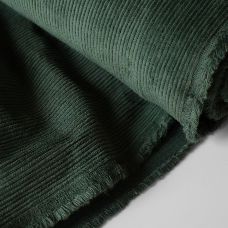 May include: Close-up of a roll of dark green corduroy fabric. The fabric has a ribbed texture and a soft, velvety appearance. The edges of the fabric are frayed, showing the raw texture of the material. The fabric is tightly rolled, with the texture clearly visible.