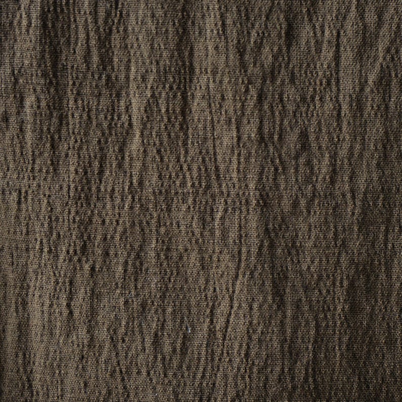 May include: Close-up of a textured, brown fabric. The fabric has a slightly wrinkled appearance, with vertical lines and subtle variations in color. The material appears to be a natural fiber, possibly linen or cotton, with a rustic aesthetic.