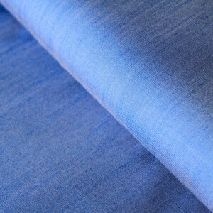 May include: Close-up of a textured, blue fabric with a gradient effect. The fabric is a medium shade of blue, with lighter shades in the folds. The weave of the fabric is visible, creating a subtle pattern.