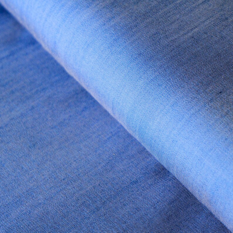 May include: Close-up of a textured, blue fabric with a gradient effect. The fabric is a medium shade of blue, with lighter shades in the folds. The weave of the fabric is visible, creating a subtle pattern.