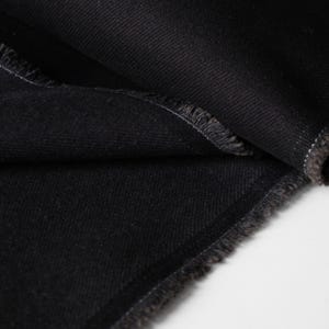 Puede incluir: Primer plano de tela vaquera negra con bordes deshilachados. La tela est&aacute; doblada, revelando la textura y el tejido del material. El color oscuro y los bordes sin rematar sugieren una prenda o un proyecto textil.