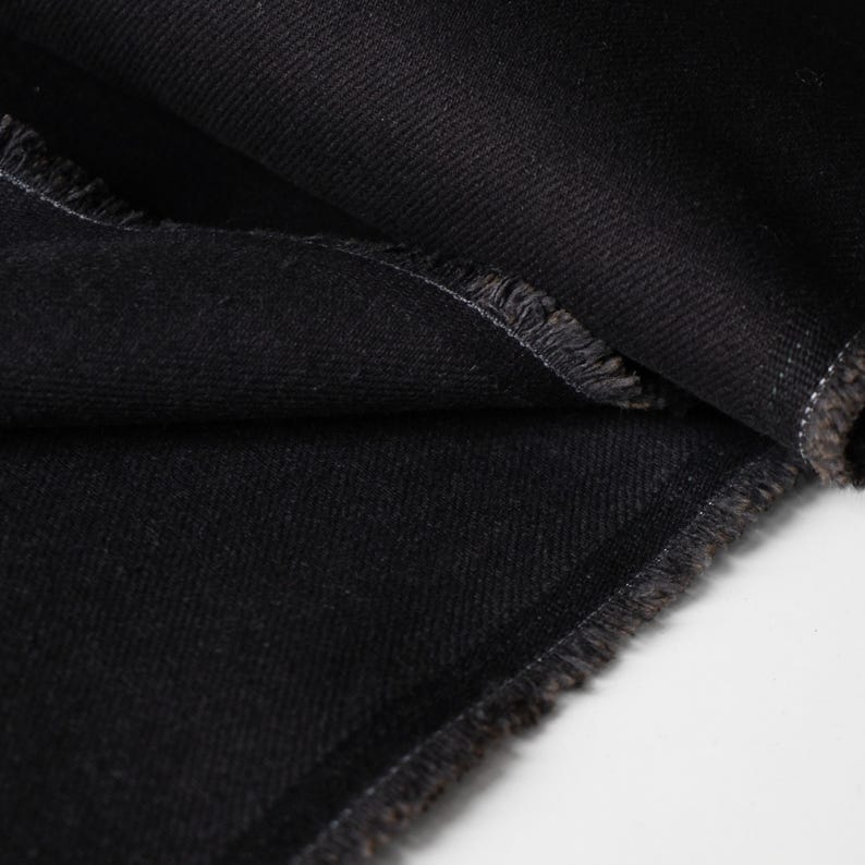 Puede incluir: Primer plano de tela vaquera negra con bordes deshilachados. La tela est&aacute; doblada, revelando la textura y el tejido del material. El color oscuro y los bordes sin rematar sugieren una prenda o un proyecto textil.