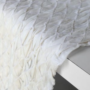May include: Close-up of a white fabric with a textured, diamond-patterned design. The fabric appears to be draped over a white surface, with the edges of the fabric visible. The texture is created by a series of gathered or pleated sections.
