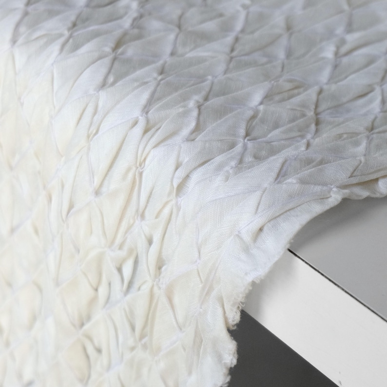 May include: Close-up of a white fabric with a textured, diamond-patterned design. The fabric appears to be draped over a white surface, with the edges of the fabric visible. The texture is created by a series of gathered or pleated sections.