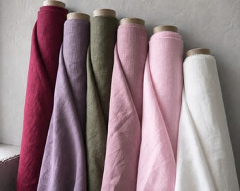 May include: Seven rolls of linen fabric in various colors, including deep red, lavender, olive green, and shades of pink and white. The fabric is tightly rolled and leans against a gray wall, showcasing the texture and color variations.