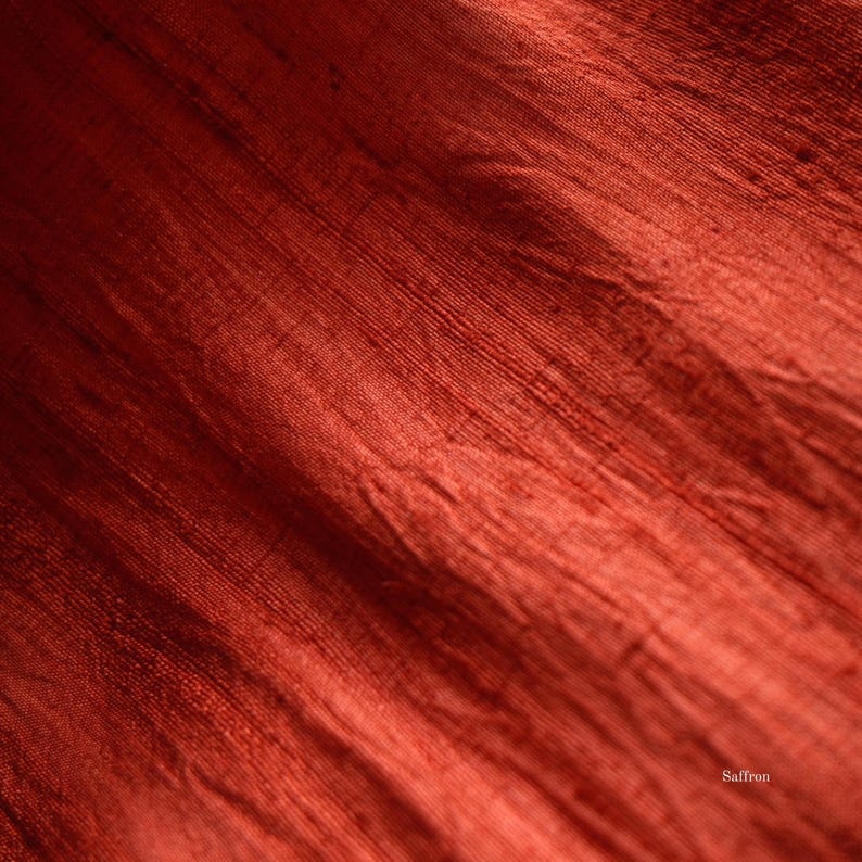May include: Close-up of a textured, rust-colored fabric with visible diagonal lines and subtle variations in tone. The fabric has a slight sheen, suggesting a silk or similar material. The word "Saffron" is visible in the bottom right corner.