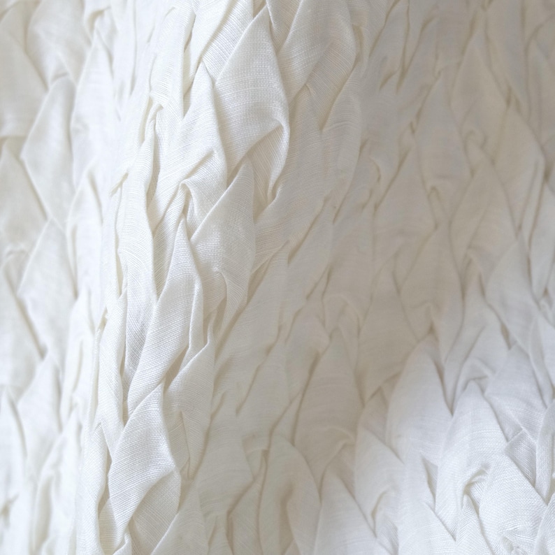 May include: Close-up of a white fabric with a textured, woven design. The fabric appears to be made of linen or a similar material, with a soft, flowing drape. The texture creates a pattern of diagonal lines.