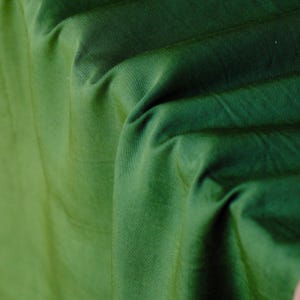 Pode incluir: Close-up de um tecido verde dobrado, mostrando textura vis&iacute;vel e varia&ccedil;&otilde;es sutis de tom. O tecido, possivelmente algod&atilde;o ou mistura, tem um leve brilho. As dobras criam sombras e destaques.