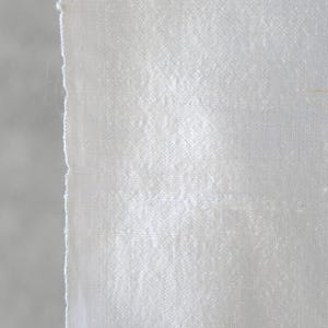 May include: Close-up of a piece of white fabric with a textured weave. The fabric has a raw, unfinished edge on the left side, revealing the individual threads. The material appears to be slightly shiny, reflecting light.