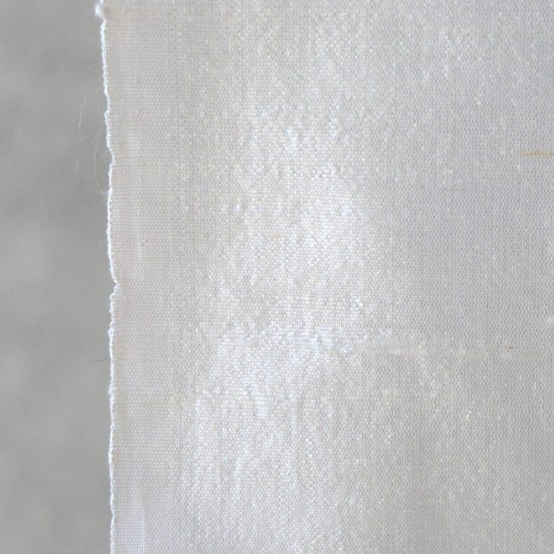 May include: Close-up of a piece of white fabric with a textured weave. The fabric has a raw, unfinished edge on the left side, revealing the individual threads. The material appears to be slightly shiny, reflecting light.