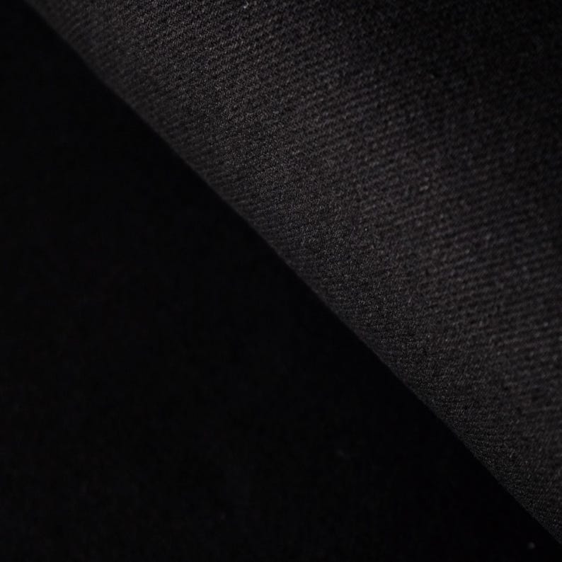 Puede incluir: Primer plano de una tela negra con una superficie texturizada. La tela est&aacute; doblada, revelando un &aacute;rea m&aacute;s oscura y sombreada. El material parece ser un tejido pesado, posiblemente mezclilla o un textil similar.