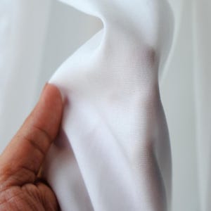 Pode incluir: Close-up de tecido branco, segurado por uma m&atilde;o. O tecido &eacute; liso e parece leve, com um ligeiro brilho. A textura do material &eacute; vis&iacute;vel, sugerindo uma trama fina. O fundo &eacute; um branco suave e desfocado.