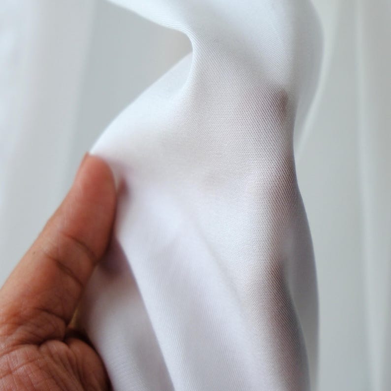 Pode incluir: Close-up de tecido branco, segurado por uma m&atilde;o. O tecido &eacute; liso e parece leve, com um ligeiro brilho. A textura do material &eacute; vis&iacute;vel, sugerindo uma trama fina. O fundo &eacute; um branco suave e desfocado.