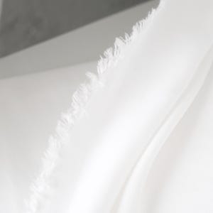 Puede incluir: Primer plano de tela blanca con borde deshilachado. La tela es lisa y parece estar drapeada, con pliegues y sombras suaves. El material es de un blanco brillante, lo que sugiere una est&eacute;tica limpia y minimalista.