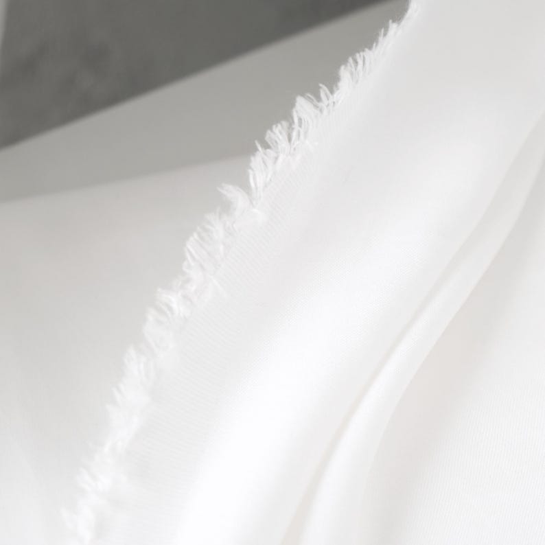 Puede incluir: Primer plano de tela blanca con borde deshilachado. La tela es lisa y parece estar drapeada, con pliegues y sombras suaves. El material es de un blanco brillante, lo que sugiere una est&eacute;tica limpia y minimalista.