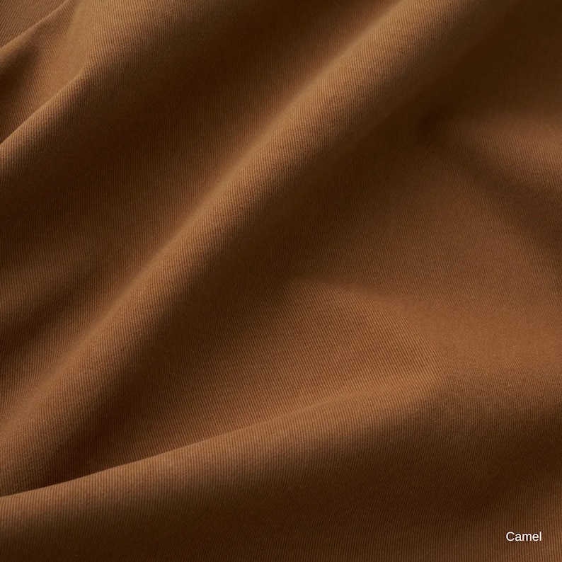 K&ouml;nnte beinhalten: Ein einfarbiger, kamelfarbener Stoff mit einer glatten, matten Textur und sichtbaren diagonalen Weblinien. Das Material ist so drapiert, dass weiche, flie&szlig;ende Falten und Schatten auf der Oberfl&auml;che entstehen. Das Wort Camel steht unten rechts.