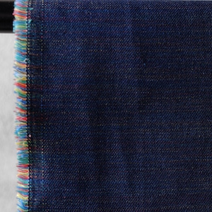 Op de afbeelding: Close-up van een donkerblauwe denimstof met een getextureerde weefsel. De rand van de stof is gerafeld en onthult kleurrijke draden van geel, rood en blauw. De stof wordt waarschijnlijk gebruikt voor kleding of woondecoratie.