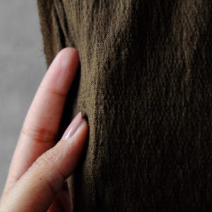 May include: Close-up of a textured, olive-green fabric. The fabric has a slightly wrinkled appearance, with visible fibers and a matte finish. The color is a deep, earthy tone, suggesting a natural material like linen or cotton.