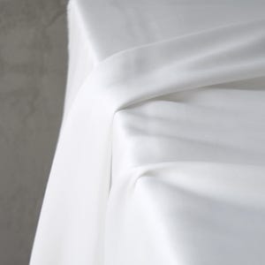 Puede incluir: Primer plano de tela blanca drapeada sobre una superficie. La tela tiene una textura suave y est&aacute; doblada en m&uacute;ltiples lugares, creando sombras y reflejos. El fondo es de color gris s&oacute;lido.