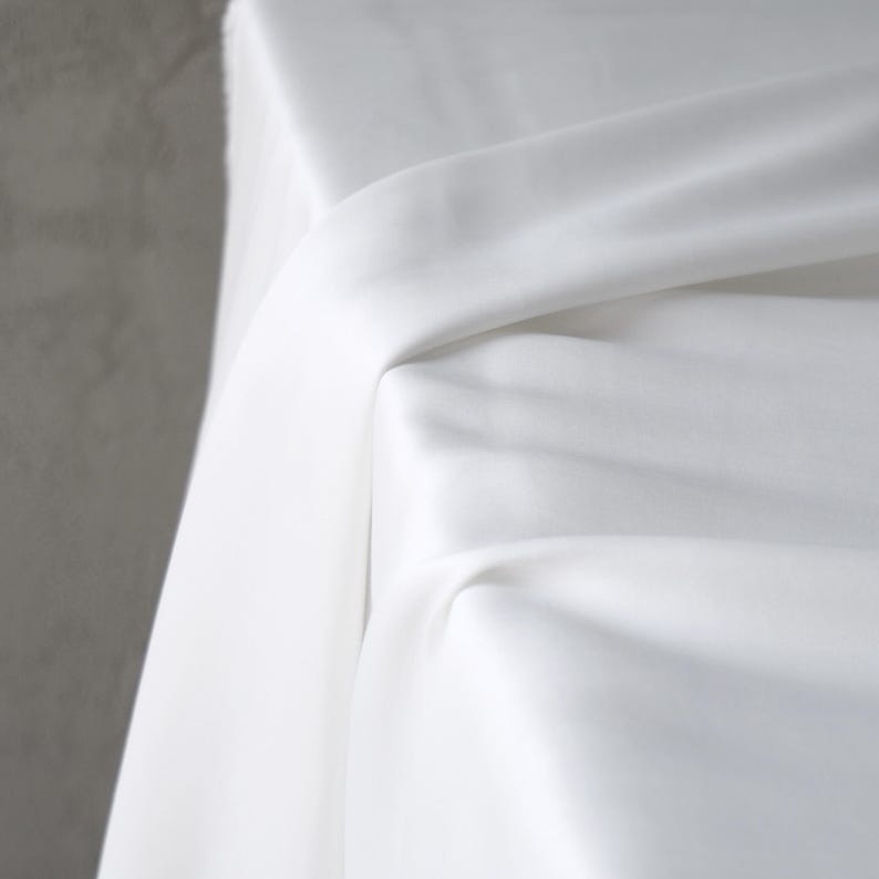 Puede incluir: Primer plano de tela blanca drapeada sobre una superficie. La tela tiene una textura suave y est&aacute; doblada en m&uacute;ltiples lugares, creando sombras y reflejos. El fondo es de color gris s&oacute;lido.