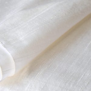 May include: Close-up of a white fabric with a subtle sheen. The fabric is folded, revealing layers and textures. The material appears to be lightweight, with visible vertical lines, suggesting a linen or similar weave.