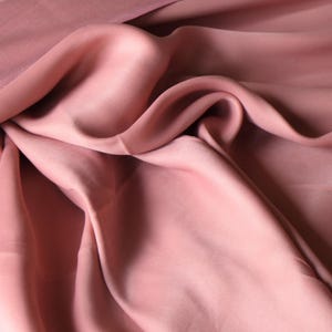Puede incluir: Primer plano de una tela rosa empolvado fluida con pliegues suaves y un brillo sutil. La tela parece ligera, posiblemente seda o un material similar, con diferentes tonos de rosa.