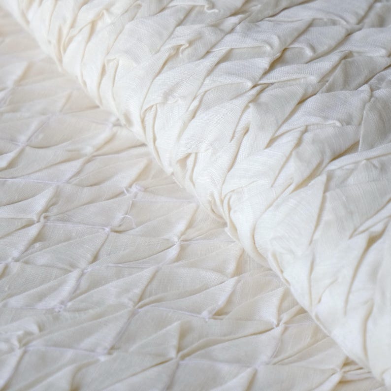 May include: Close-up of a cream-colored fabric with a textured, quilted design. The fabric is folded, revealing the intricate pattern of raised, diamond-shaped sections. The material appears soft and possibly linen or cotton.