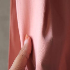 Puede incluir: Primer plano de una secci&oacute;n de tela rosa. La tela es lisa y parece ser de color liso. Un dedo toca la tela, mostrando la textura y las variaciones de color.