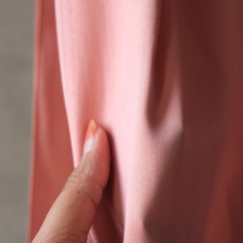 Puede incluir: Primer plano de una secci&oacute;n de tela rosa. La tela es lisa y parece ser de color liso. Un dedo toca la tela, mostrando la textura y las variaciones de color.