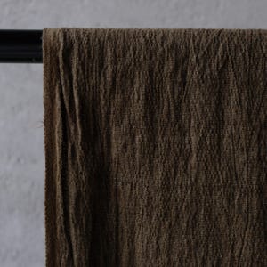 May include: Close-up of a brown fabric draped over a black rod. The fabric has a textured, wrinkled appearance, with visible vertical lines. The color is a deep, earthy brown, suggesting a natural material like linen or cotton.