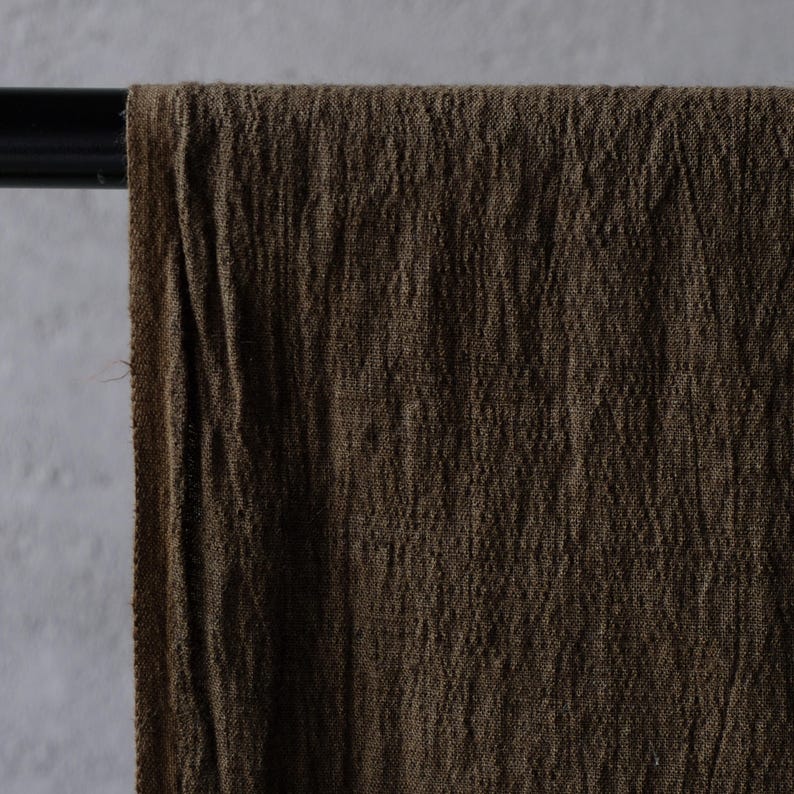 May include: Close-up of a brown fabric draped over a black rod. The fabric has a textured, wrinkled appearance, with visible vertical lines. The color is a deep, earthy brown, suggesting a natural material like linen or cotton.