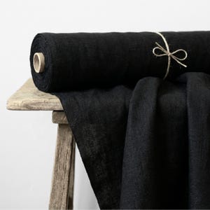 May include: A roll of black fabric sits on a rustic wooden table. The fabric is dark and appears to be linen or a similar material. A length of the fabric is draped over the table's edge. The roll is tied with twine.