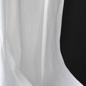 May include: Close-up of white fabric with soft folds and a textured appearance. The fabric is draped, creating shadows and highlights against a black background. The material appears to be lightweight and flowing.