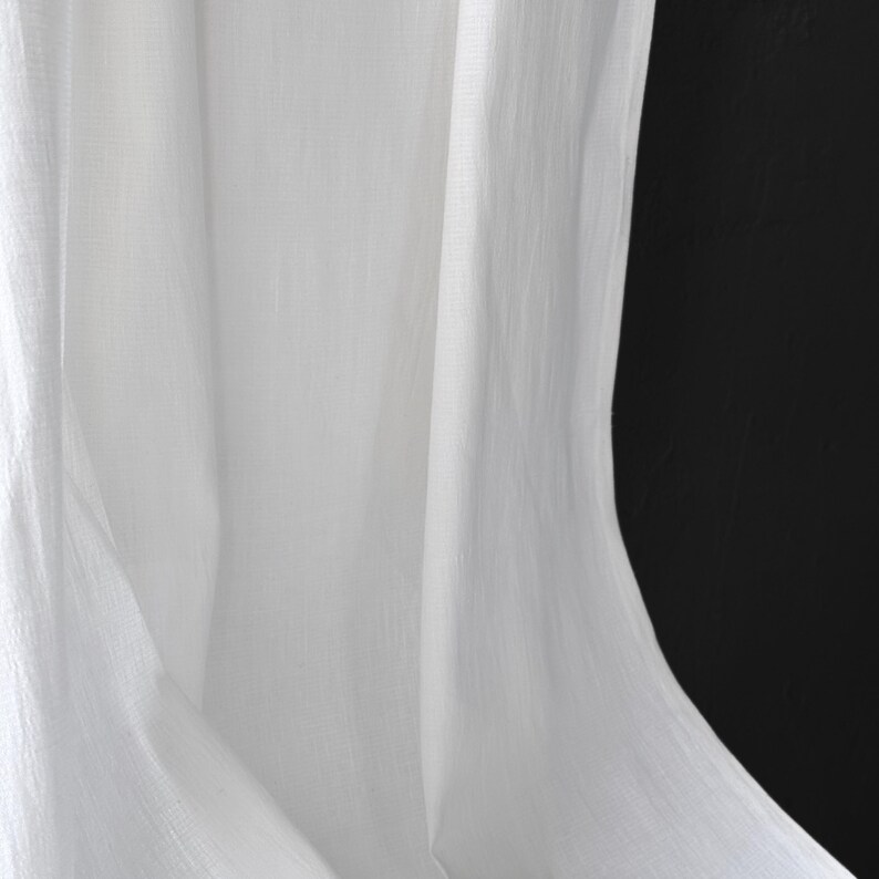 May include: Close-up of white fabric with soft folds and a textured appearance. The fabric is draped, creating shadows and highlights against a black background. The material appears to be lightweight and flowing.